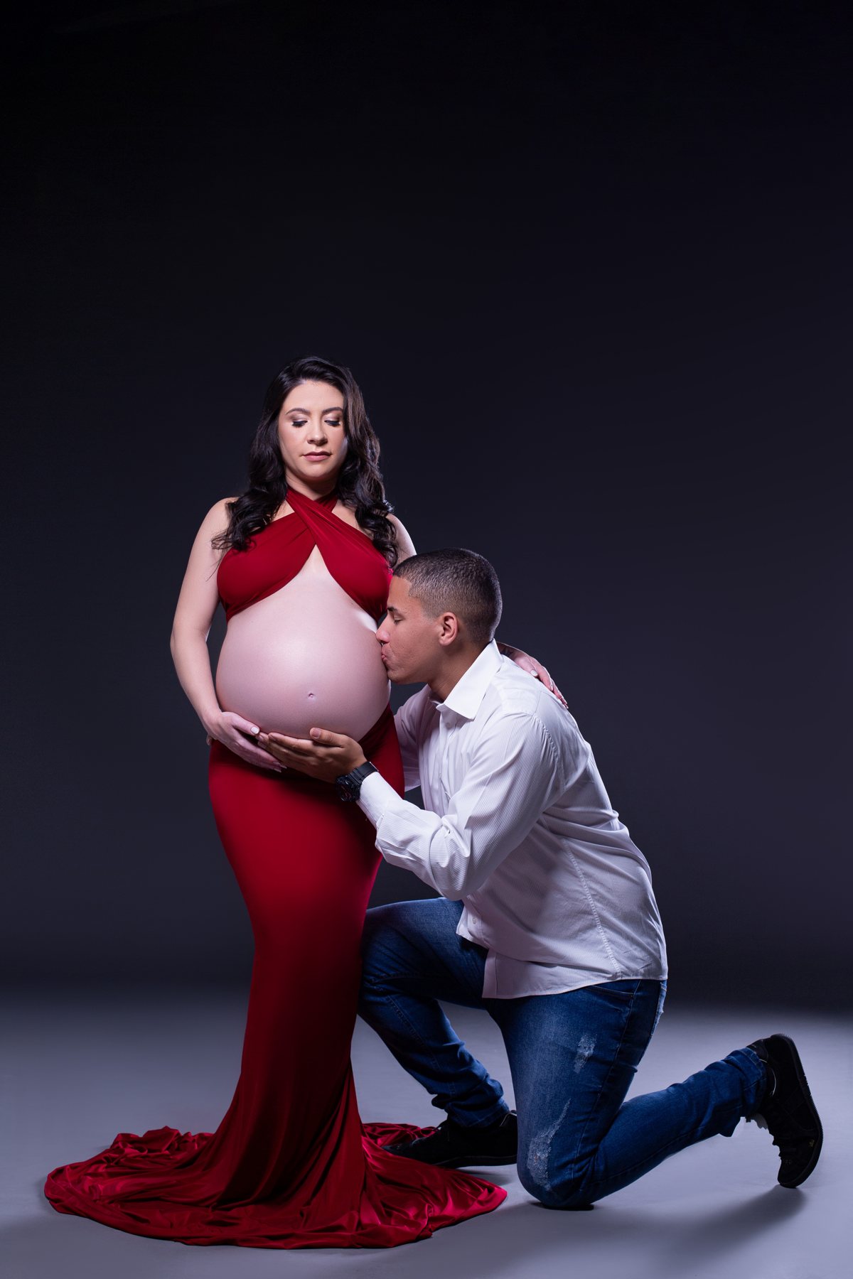 Priscilla de vestido vermelho e Henrique ajoelhado ao seu lado em ensaio de gestação